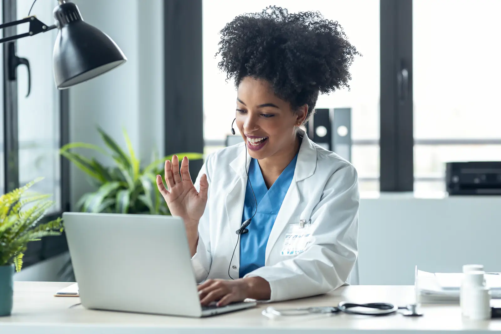 A woman in a white coat and headset smiles and waves at a laptop on her desk.