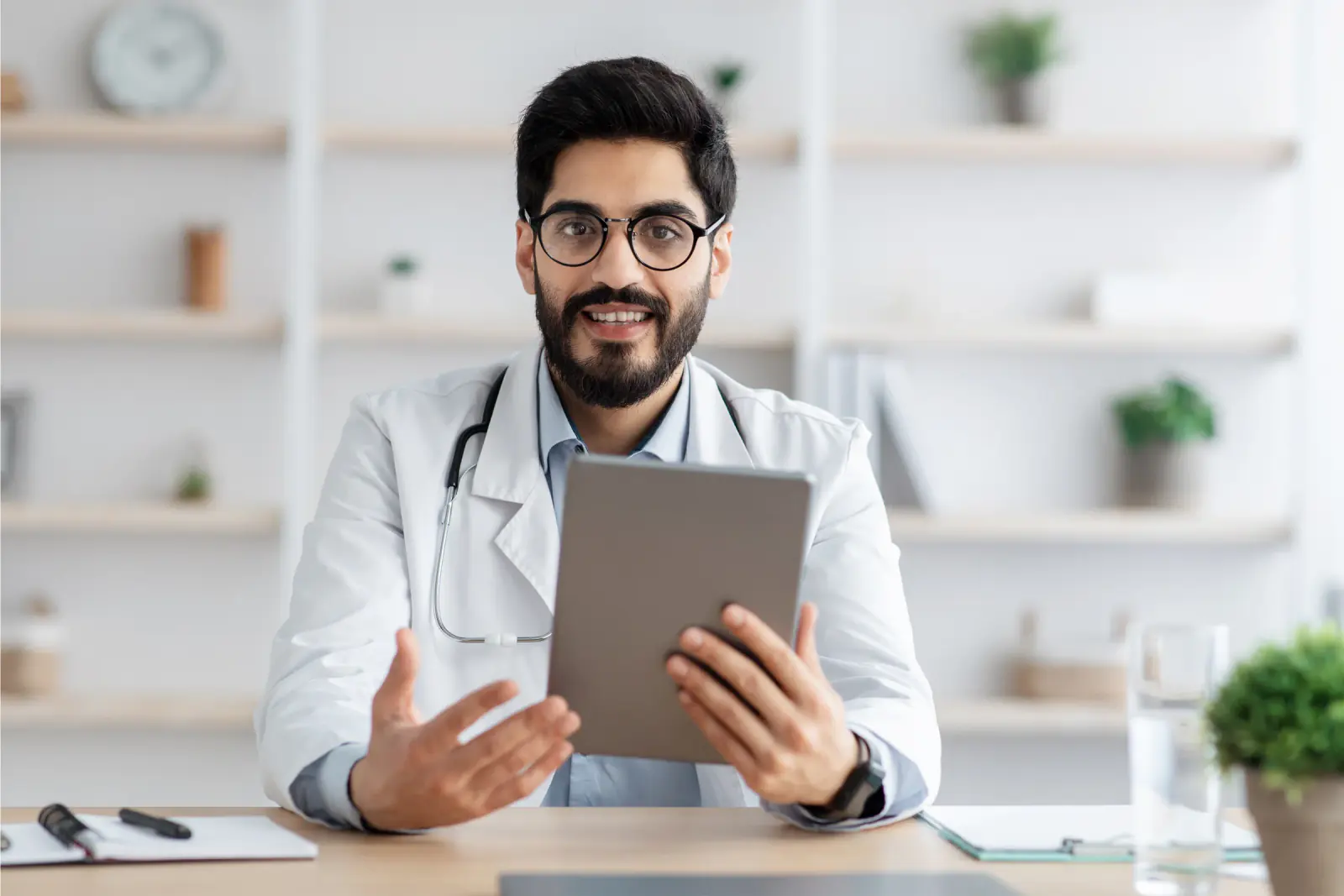 A man in glasses and a white coat holds a tablet and talks toward the camera.
