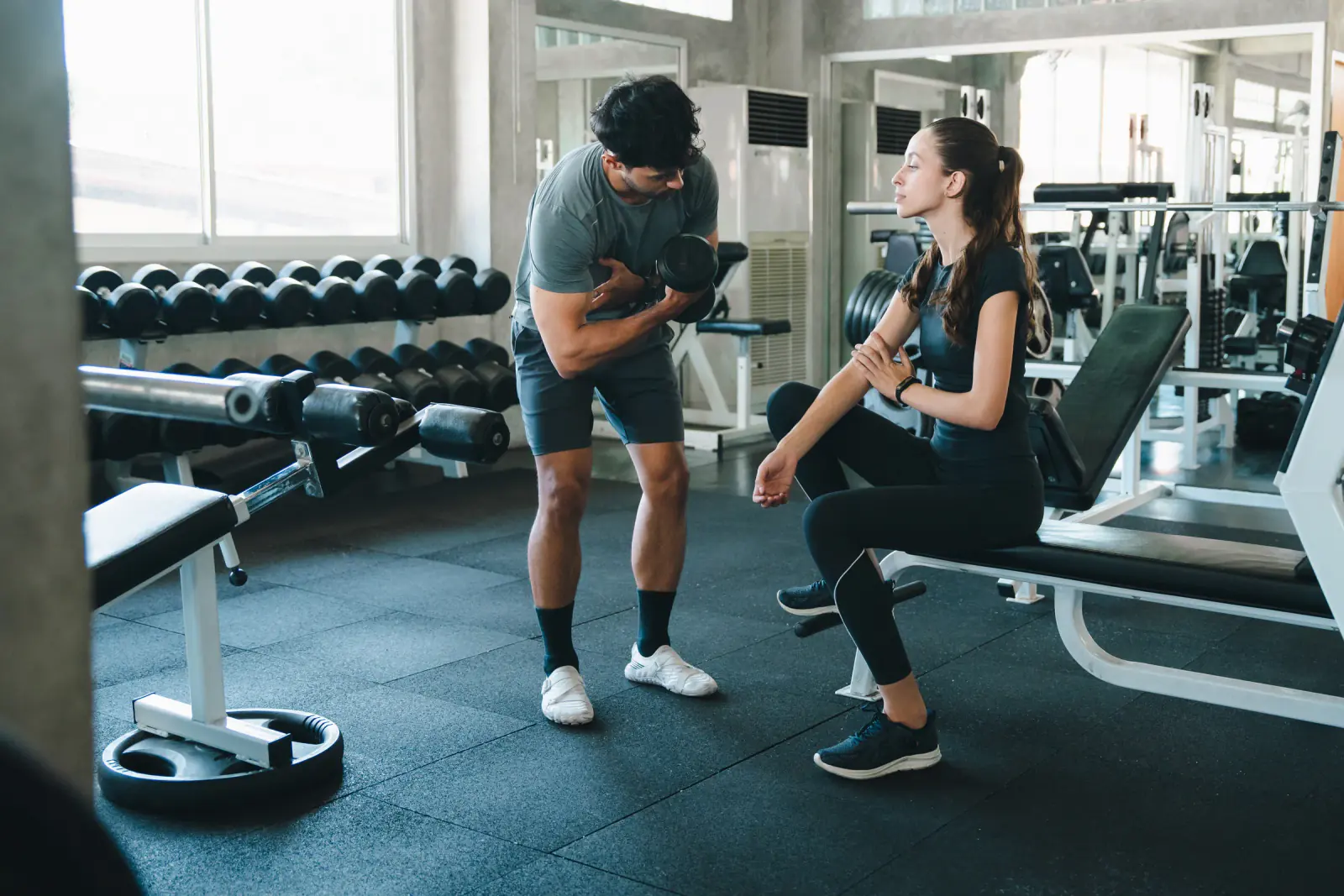 A trainer demonstrates a dumbbell curl to a woman seated on a gym bench.