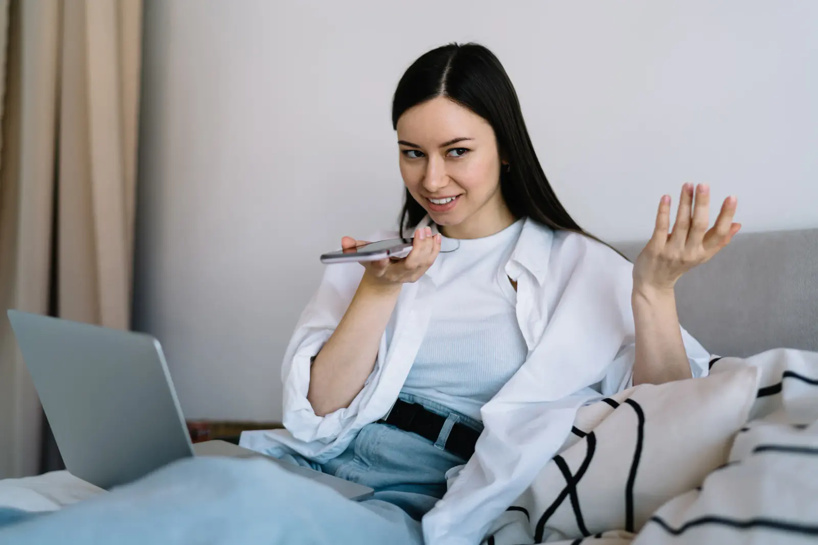 A woman sitting on a sofa with a laptop talks into her phone and smiles.