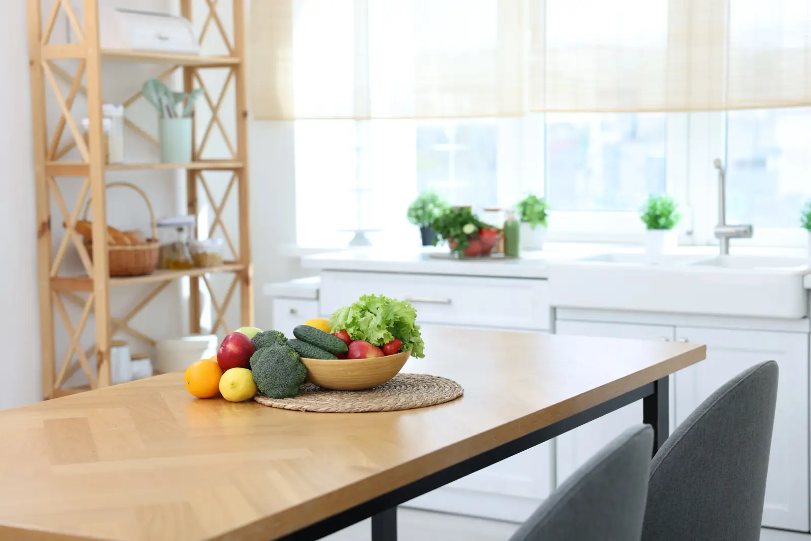 A bowl of fruit and vegetables on a wooden table in a bright kitchen.